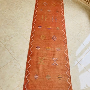 May include: A long, rectangular rug with a rust-colored background and a geometric pattern in shades of orange, yellow, purple, and white. The rug has a fringe on the bottom edge.