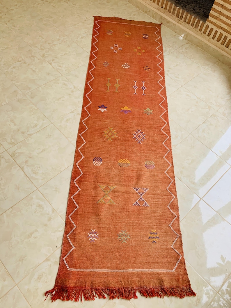 May include: A long, rectangular rug with a rust-colored background and a geometric pattern in shades of orange, yellow, purple, and white. The rug has a fringe on the bottom edge.