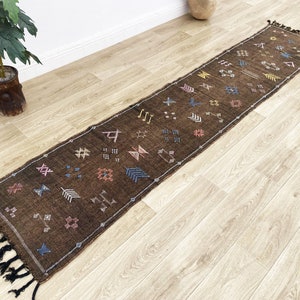 以下が含まれることがあります： ピンク、ブルー、イエロー、ホワイトの幾何学模様が施されたブラウンのランナーラグ。ラグの両端には黒いフリンジが付いています。