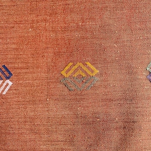 May include: A close-up of a woven rug with a geometric pattern in shades of blue, yellow, and brown. The rug has a textured surface and a warm, earthy tone.