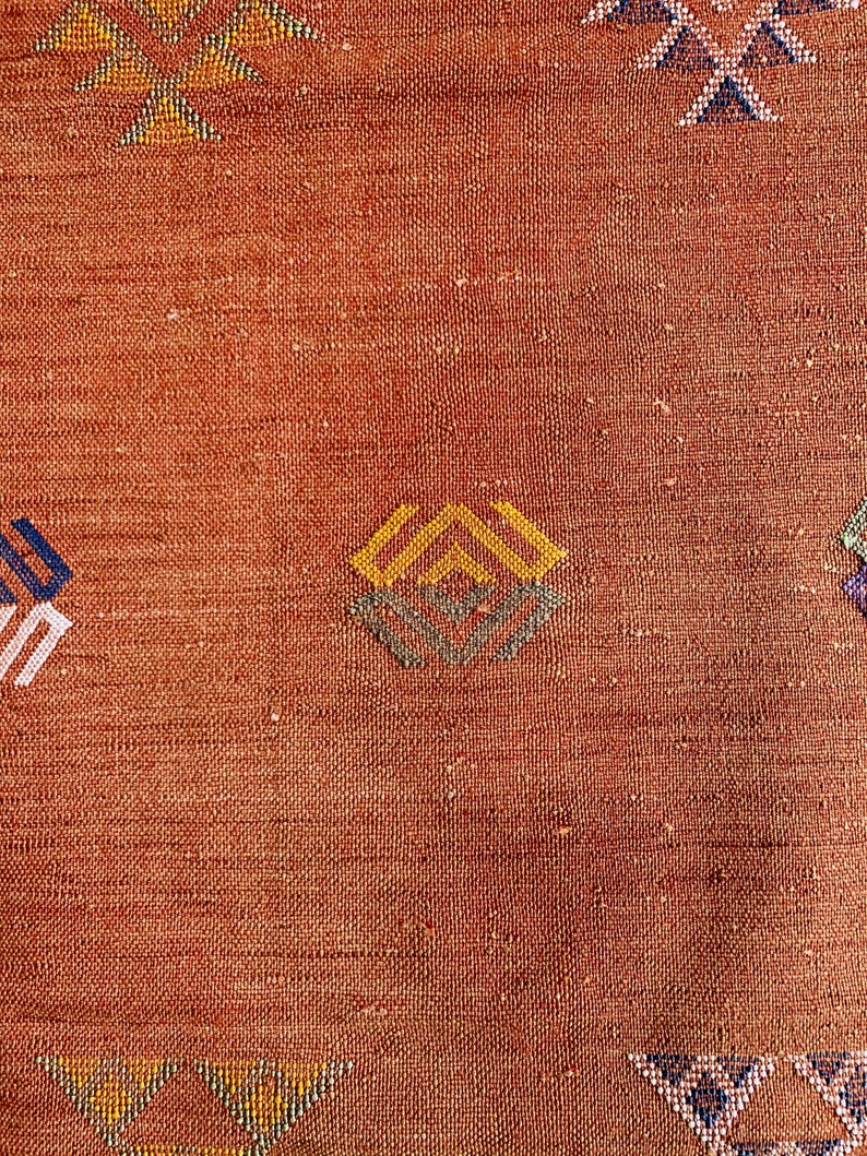 May include: A close-up of a woven rug with a geometric pattern in shades of blue, yellow, and brown. The rug has a textured surface and a warm, earthy tone.