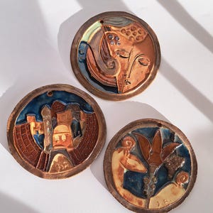 Könnte beinhalten: Drei runde Keramikkunstwerke mit detaillierten Reliefdesigns. Jedes Stück zeigt eine andere Szene: ein Gesicht, eine Stadtlandschaft und eine Figur mit Pflanzen. Die Stücke haben einen bronzefarbenen Rand und farbige Akzente.