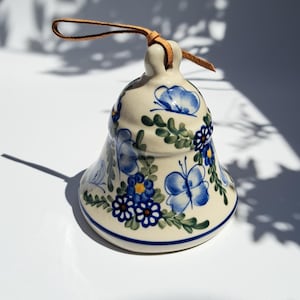 Op de afbeelding: Een keramische bel met een crèmekleurige basis, versierd met blauwe bloemen- en vlindermotieven. De bel heeft een bruine leren lus om op te hangen. Het ontwerp omvat blauwe bloemen, vlinders en groene bladeren. De bel is ongeveer 10 cm hoog.