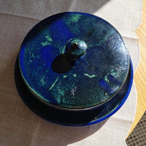 Op de afbeelding: Een blauwe keramische schaal met een deksel. Het deksel heeft een groene en blauwe glazuur met een knop bovenop. De schaal staat op een wit linnen tafelkleed.