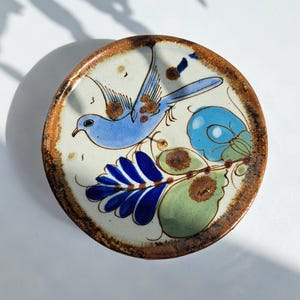 Op de afbeelding: Een ronde keramische schaal met een crèmekleurige basis en een bruine rand. Het oppervlak is voorzien van een handgeschilderd ontwerp van een lichtblauwe vogel in vlucht, vergezeld door gestileerde groene en blauwe bladeren en bloemmotieven met bruine spikkels.