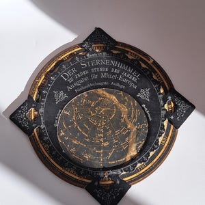 Könnte beinhalten: Eine Sternenkarte im Vintage-Stil in Schwarz und Gold. Die Karte zeigt eine detaillierte Sternenkarte in der Mitte, umgeben von deutschem Text. Das Design umfasst dekorative Elemente und eine einzigartige Form.