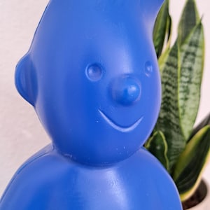 Könnte beinhalten: Eine einfarbige blaue Figur eines lächelnden Zeichentrickcharakters mit stacheligen Haaren. Die Figur hat eine runde Nase, kleine Augen und einen fröhlichen Ausdruck. Eine Topfpflanze mit grünen und gelben Blättern befindet sich im Hintergrund.