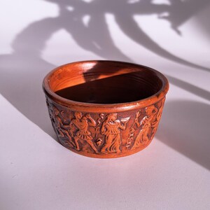 Op de afbeelding: Een ronde, terracotta-kleurige kom met een gedetailleerd reliëf van figuren in klassieke kleding. De buitenkant van de kom heeft een herhalend friesontwerp. De binnenkant is een donkerdere bruintint. De kom heeft een diameter van ongeveer 10 cm.