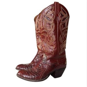 May include: A pair of dark brown cowboy boots featuring a textured, scale-like pattern on the foot and a smooth leather shaft. The upper shaft is decorated with intricate, light-colored stitching in a floral-inspired design and pull tabs.