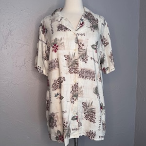 May include: Cream-colored button-down shirt with a tropical floral and animal print. The short-sleeved shirt features a collar and a series of small buttons down the front. The print includes maroon flowers, brown leopard spots, and green leaves.