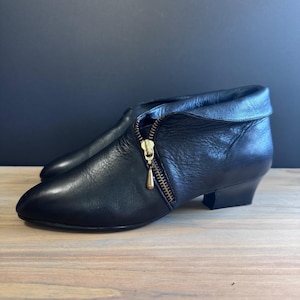 May include: Black leather ankle boots with a gold-toned zipper and a small heel. The boots have a rounded toe and a folded-over cuff. The zipper pull has a small, decorative charm.
