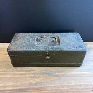 May include: An olive green metal toolbox with a handle and a lock. The box has a rectangular shape and shows signs of wear, with some rust and discoloration. The handle is a curved metal bar. The box is on a wooden surface.