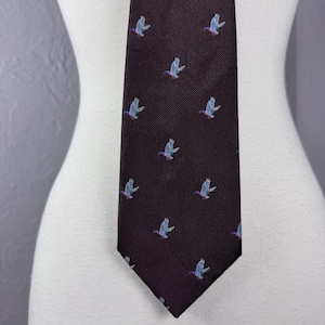 May include: A dark brown necktie featuring a repeating pattern of small, embroidered flying birds. The birds are depicted in shades of light blue, white, and red, set against a textured, diagonal weave fabric that runs throughout the entire tie.