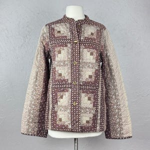 May include: A quilted jacket with a stand-up collar and long sleeves. The jacket features a patchwork design in shades of brown, beige, and cream, with gold-colored buttons down the front. The jacket is displayed on a mannequin.