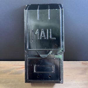 May include: A black metal mailbox with the word "MAIL" in white lettering. The mailbox has a rectangular shape with a hinged door at the bottom and a slot at the top. The mailbox is mounted on a wooden surface.