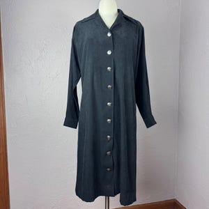 May include: A long-sleeved, charcoal black button-down dress featuring a collared neckline and a vertical row of nine circular silver-toned metal buttons down the center front. The garment has a soft, matte texture and a straight silhouette.