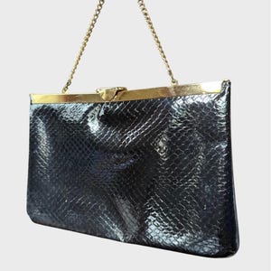 May include: Black clutch with a gold-tone clasp and chain strap. The rectangular bag has a textured, snake-like pattern. The gold chain is attached to the top of the bag, and the clasp is also gold.