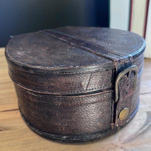 May include: A vintage, round, brown leather hat box with a strap and buckle closure. The box has a distressed appearance, suggesting age and use. The lid and base are stitched together, and the box sits on a wooden surface.