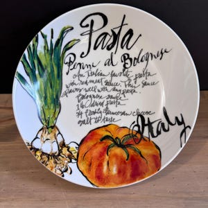 May include: White ceramic pasta bowl with a hand-painted design. The bowl features the word "Pasta" in black script, along with a recipe for Penne al Bolognese. Illustrations of a tomato and onion are also included. The word "Italy" is written on the bowl.