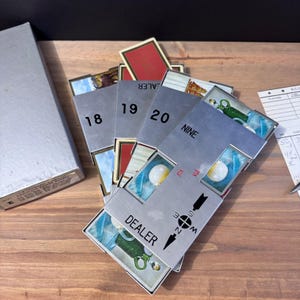May include: A vintage card game with silver card holders, numbered 18, 19, and 20. The cards feature illustrations and the word "DEALER" on one holder. A silver box and score sheet are also visible.