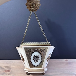 May include: Cream-colored hanging planter with gold accents and a floral design. The planter is suspended by a gold chain from a decorative ceiling mount. The planter has a rectangular shape with a decorative gold trim.