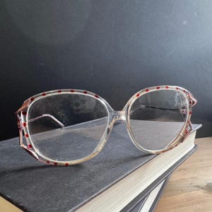 May include: Clear plastic eyeglasses featuring a rounded frame design with small, evenly spaced red dots along the outer rim. The frames include pointed, wing-like accents at the outer corners and thin, matching clear plastic temples.