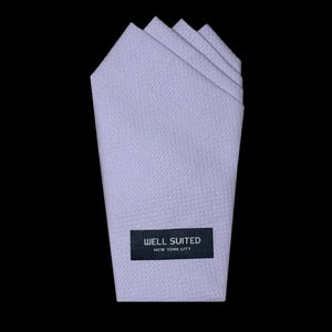 May include: A light lavender pocket square with a subtle, repeating geometric honeycomb texture. The fabric is folded into four distinct points at the top. A rectangular black label is stitched near the bottom, featuring the white text WELL SUITED NEW YORK CITY.