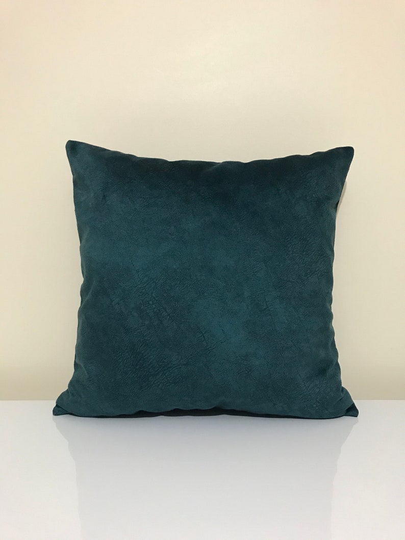 Op de afbeelding: Een vierkant decoratief kussen in teal. Het kussen heeft een getextureerde, su&egrave;de-achtige uitstraling. Het kussen ligt op een wit oppervlak. Een item voor in huis.