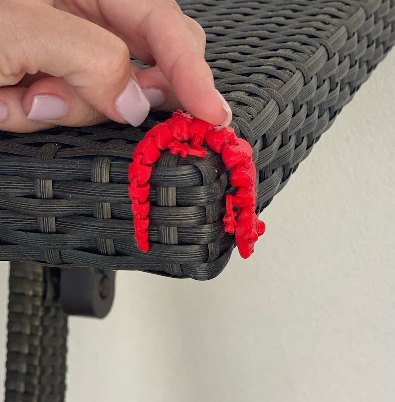 May include: A red 3D printed gecko figurine is perched on the edge of a black wicker table.