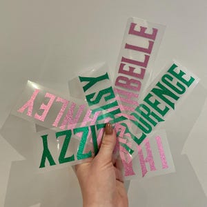 May include: A selection of heat transfer vinyl sheets featuring names in pink and green glitter. Names include Izzy, Finley, Belle, Florence, and Hi. The sheets are held in a hand against a white backdrop.