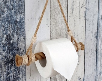 May include: A rustic toilet paper holder made of natural rope. The holder is suspended from a bronze hook and features a roll of white toilet paper. The rope is knotted at various points for a decorative touch.