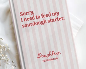 May include: A pink and white striped sourdough feeding log book with the text "Sorry, I need to feed my sourdough starter." The book also has the word "Doughlene" and "Feeding Log" printed on the cover. The book is on a white surface.
