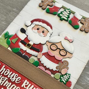 Puede incluir: Un letrero de madera con tem&aacute;tica navide&ntilde;a que presenta a Pap&aacute; Noel y Mam&aacute; Noel. El letrero tiene un borde rojo y marr&oacute;n con el texto "This house runs on Christmas and..." Decorado con hombres de jengibre, calcetines y &aacute;rboles de Navidad.