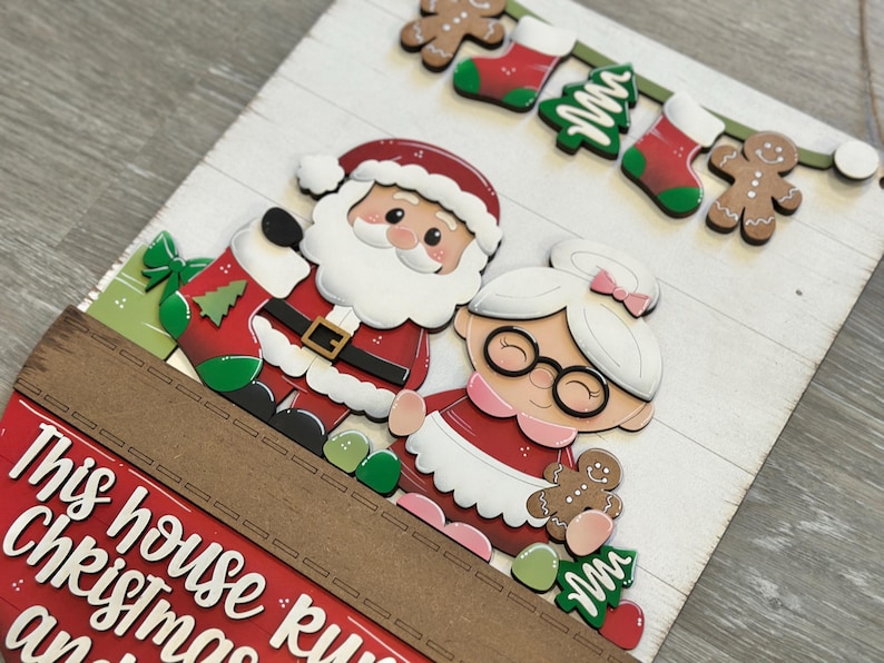 Puede incluir: Un letrero de madera con tem&aacute;tica navide&ntilde;a que presenta a Pap&aacute; Noel y Mam&aacute; Noel. El letrero tiene un borde rojo y marr&oacute;n con el texto "This house runs on Christmas and..." Decorado con hombres de jengibre, calcetines y &aacute;rboles de Navidad.