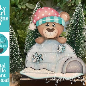 May include: A wooden Christmas decoration featuring a bear wearing a pink and teal hat, peeking over an igloo. The igloo has snowflake accents. The text "Lucky Heart Designs Co" and "Digital cut file instant download" are visible.