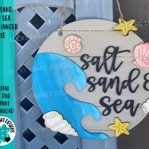 May include: A round wooden door hanger with the text "salt sand & sea" in black lettering. The background is painted in a light gray color with a blue wave design. The door hanger is decorated with seashells, starfish, and a seashell in pink, yellow, and white.