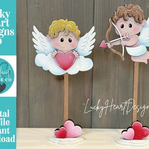 May include: Two Valentine's Day decorations featuring cherubs. One cherub holds a heart, the other aims a bow and arrow. Both have wings and are mounted on wooden sticks with heart-shaped bases. The image includes the text "Lucky Heart Designs Co."