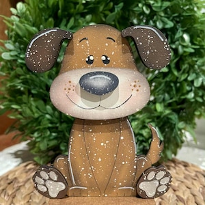 May include: A wooden dog figurine with a brown body and head, featuring a black nose, white accents, and a smiling expression. The dog is sitting and has a small tail. The figurine is on a black base and has white speckles.