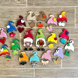 Op de afbeelding: Een verzameling kleurrijke houten dwerg ornamenten met verschillende thema's, waaronder harten, klavertjesvier, regenbogen, pompoenen en watermeloenen. De dwergen zijn geschilderd in felle kleuren en hebben verschillende gezichtsuitdrukkingen.