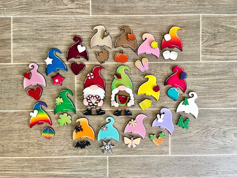 Op de afbeelding: Een verzameling kleurrijke houten dwerg ornamenten met verschillende thema's, waaronder harten, klavertjesvier, regenbogen, pompoenen en watermeloenen. De dwergen zijn geschilderd in felle kleuren en hebben verschillende gezichtsuitdrukkingen.