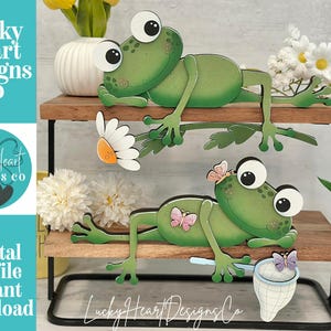 Puede incluir: Dos recortes de ranas de madera verde con ojos grandes se exhiben en un estante de varios niveles. Una rana sostiene una margarita, la otra una mariposa. El estante también contiene flores y el texto "Lucky Heart Designs Co" y "Digital cut file instant download".