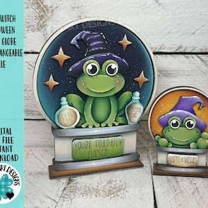 May include: Two wooden Halloween snow globes featuring a green frog witch wearing a purple hat. The larger globe has the text "You're Toadally Cursed" and the smaller globe has "Warts & Wishes". The background is a starry night.