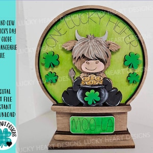 May include: A wooden snow globe with a Highland cow holding a pot of gold. The globe is decorated with shamrocks and the text "One Lucky Heifer". The base of the globe has the text "Moo-La".