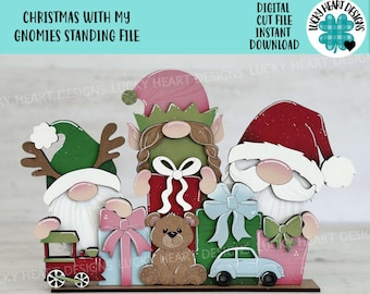 May include: A Christmas-themed wooden craft project featuring three gnomes standing in front of presents. The gnomes are wearing red, green, and white hats and have white beards. The presents are wrapped in red, green, blue, and pink paper with bows. There is a toy train, a teddy bear, and a blue toy car in front of the gnomes.