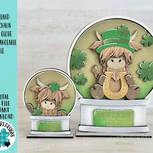 May include: Two wooden snow globes featuring a Highland cow wearing a green hat and holding a horseshoe. The smaller globe reads "Moo-la" and the larger globe reads "One lucky heifer".