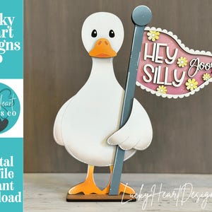 Könnte beinhalten: Eine weiße Gänsefigur mit orangefarbenem Schnabel und Füßen, die einen grauen Stab mit einem rosa Wimpel hält. Die Flagge trägt den weißen Text "HEY SILLY GOOSE" und ist mit kleinen gelben Blumen verziert. Das Stück steht auf einem flachen Sockel.