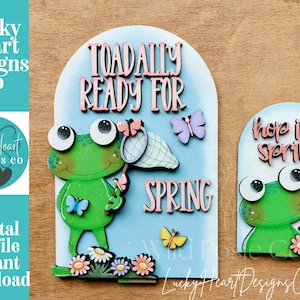 Peut inclure: Décorations printanières en bois avec une grenouille verte, des papillons et des fleurs. Un panneau indique "Toadally Ready For Spring", l'autre "Hop Into Spring". Fond bleu clair.
