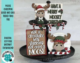 May include: A three-tiered tray decorated for Christmas with a moose theme. The top tier features a wooden moose with a red Santa hat and a green bow. The middle tier has a sign that reads "Have a Merry Moosey Christmas". The bottom tier has a sign that reads "Not a creature was stirring not even a moose".