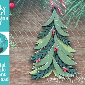May include: A layered wooden Christmas tree ornament featuring shades of light and dark green foliage with small red berries. The tree has a speckled white finish on the leaves and is suspended by a red and white twisted string at the top.