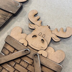 May include: A wooden moose cutout with a smiling face, holding a heart in its hooves, is sitting inside a wooden basket with a checkered pattern.